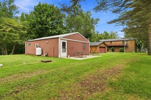 En röd metallbod med dörr och fönster, ett picknickbord och en gunga på en gräsig gård med träd i bakgrunden.