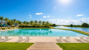 A swimming pool with lounge chairs and a wooden deck, surrounded by palm trees and a view of a lake. - Luxury Rooftop with Views of Cana: Pool, Sunset, Golf (Punta Cana)