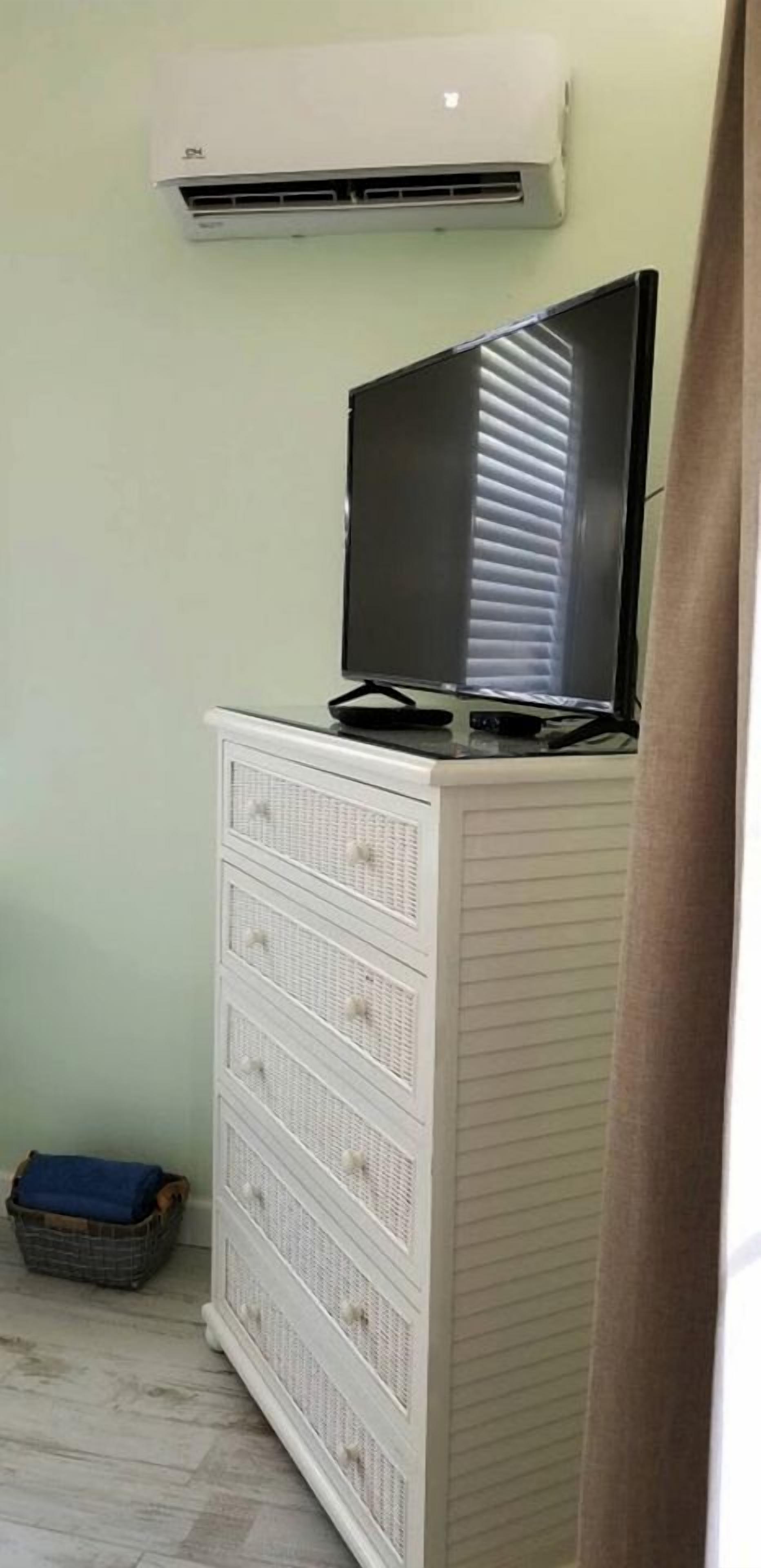 A room with a white dresser, a television on top, and an air conditioning unit on the wall.