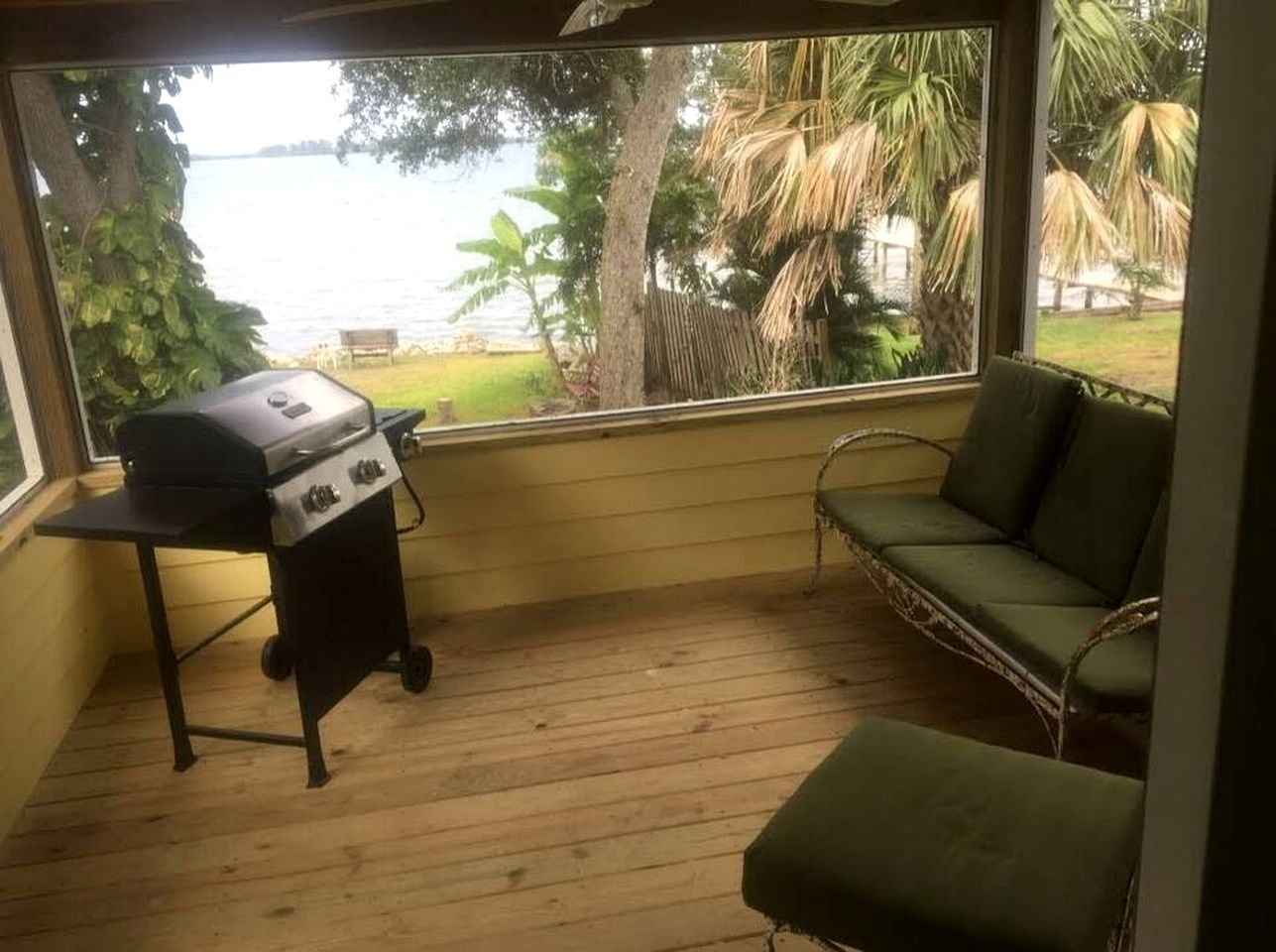 A covered outdoor area with a grill, a green cushioned seating area, and a view of a body of water and greenery.
