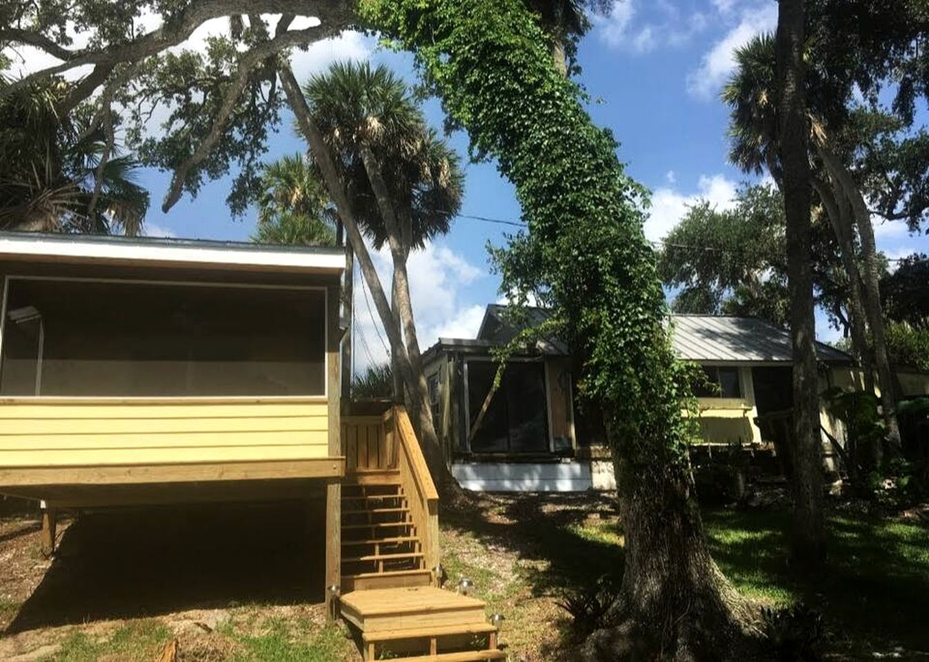 Casa móvel com escada de madeira para um deck, cercada por árvores e céu claro.