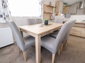 A dining area with a wooden table and grey upholstered chairs, a potted plant, and a kitchen in the background. - Rosella 38 (Morecambe)