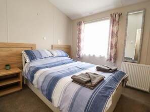 A single-bed room with a wooden headboard, a bedside table with a plant, a window with curtains, and a radiator. - Rosella 38 (Morecambe)