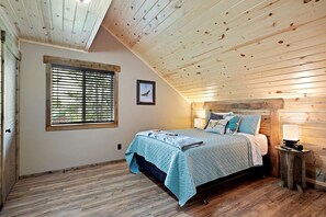 Ein Schlafzimmer mit schrägen Wänden, einem Bett mit blauer Bettdecke, einem Nachttisch mit Lampe und einem Fenster mit Jalousien.