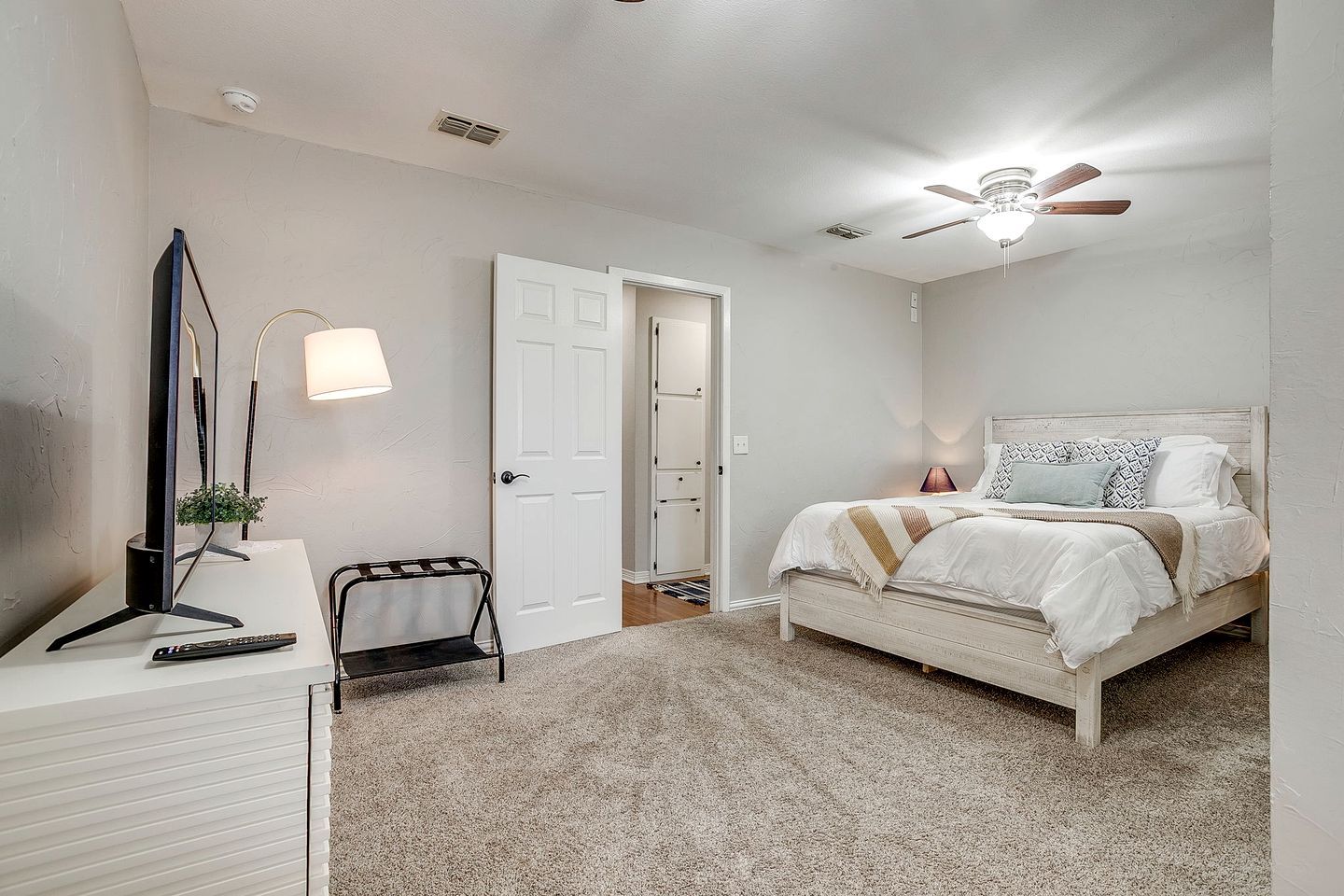 Une chambre à coucher comprenant un lit, une table de chevet, une lampe, un ventilateur de plafond et une porte donnant sur une autre pièce.