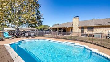 Une piscine entourée d’une terrasse délimitée par une clôture, avec des chaises longues et un espace patio couvert.