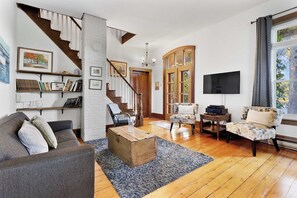 A spacious living room with a staircase, wooden flooring, a grey sofa, a patterned armchair, and a flat-screen TV mounted on the wall.