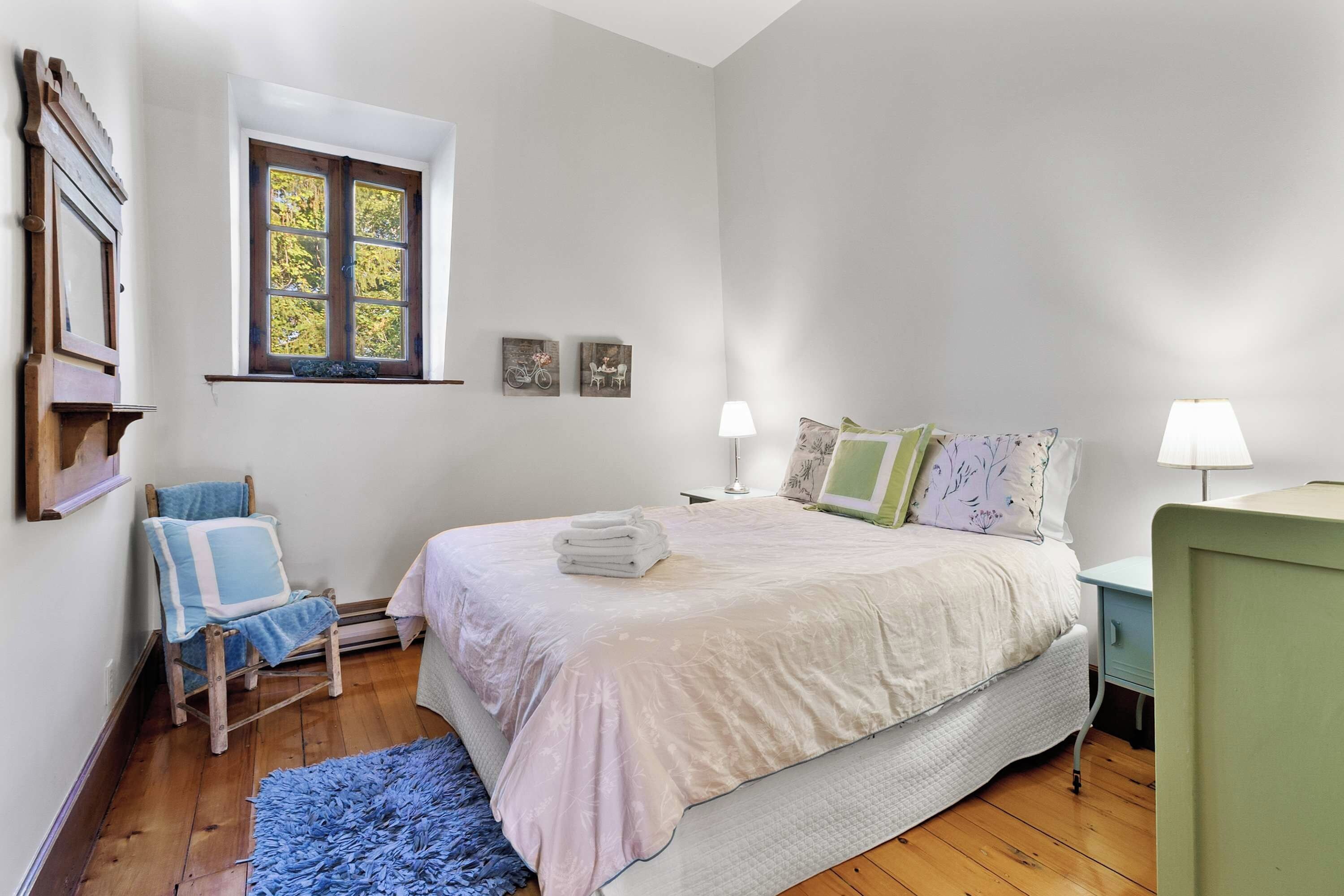 A bedroom with a bed, a chair, a window, and a nightstand.