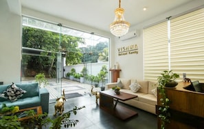 A modern living room with a sofa, coffee table, and a chandelier. There is a large window with a view of greenery outside. - Hotel Vishwa Retreat - Kandy (Kandy)