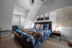 Quarto com teto inclinado, cama com colcha xadrez azul e branco, cabeceira de madeira e mesa de cabeceira com abajur.