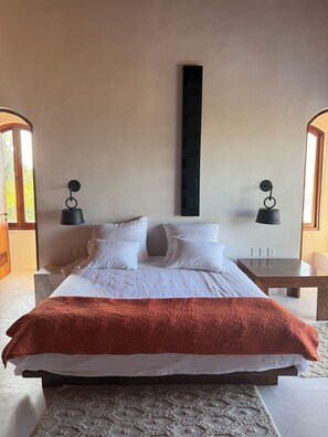 Ein modernes Schlafzimmer mit einem großen Bett, zwei Nachttischen, einer Wandleuchte und einem Blick durch einen Rundbogen.