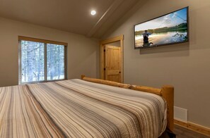 Ein Schlafzimmer mit einem großen Bett, einer Holztür, einem Fenster mit Jalousien und einem an der Wand befestigten Fernseher, der eine Anglerszene zeigt.