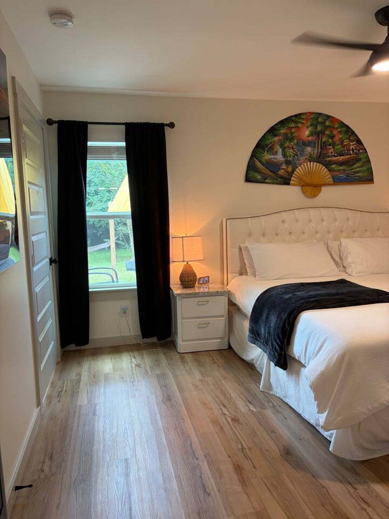 Une chambre à coucher comprenant un lit, des tables de chevet, une commode, une fenêtre avec des rideaux, un ventilateur de plafond et une œuvre d’art accrochée au mur.