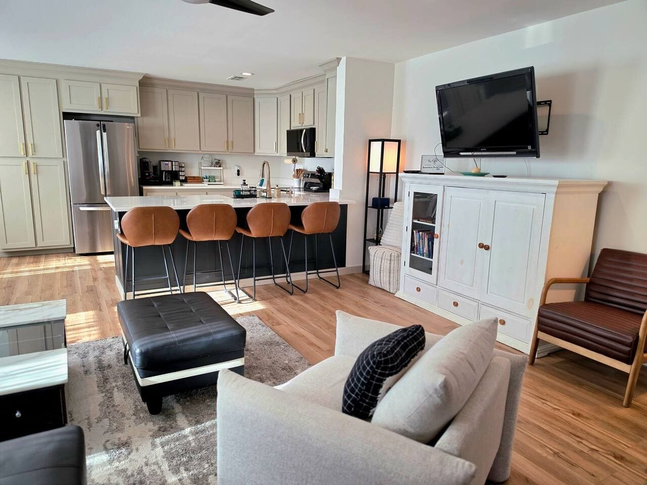 A modern living room with a sofa, ottoman, and chairs, a kitchen with a refrigerator, stove, and bar stools, and a TV mounted above a fireplace.