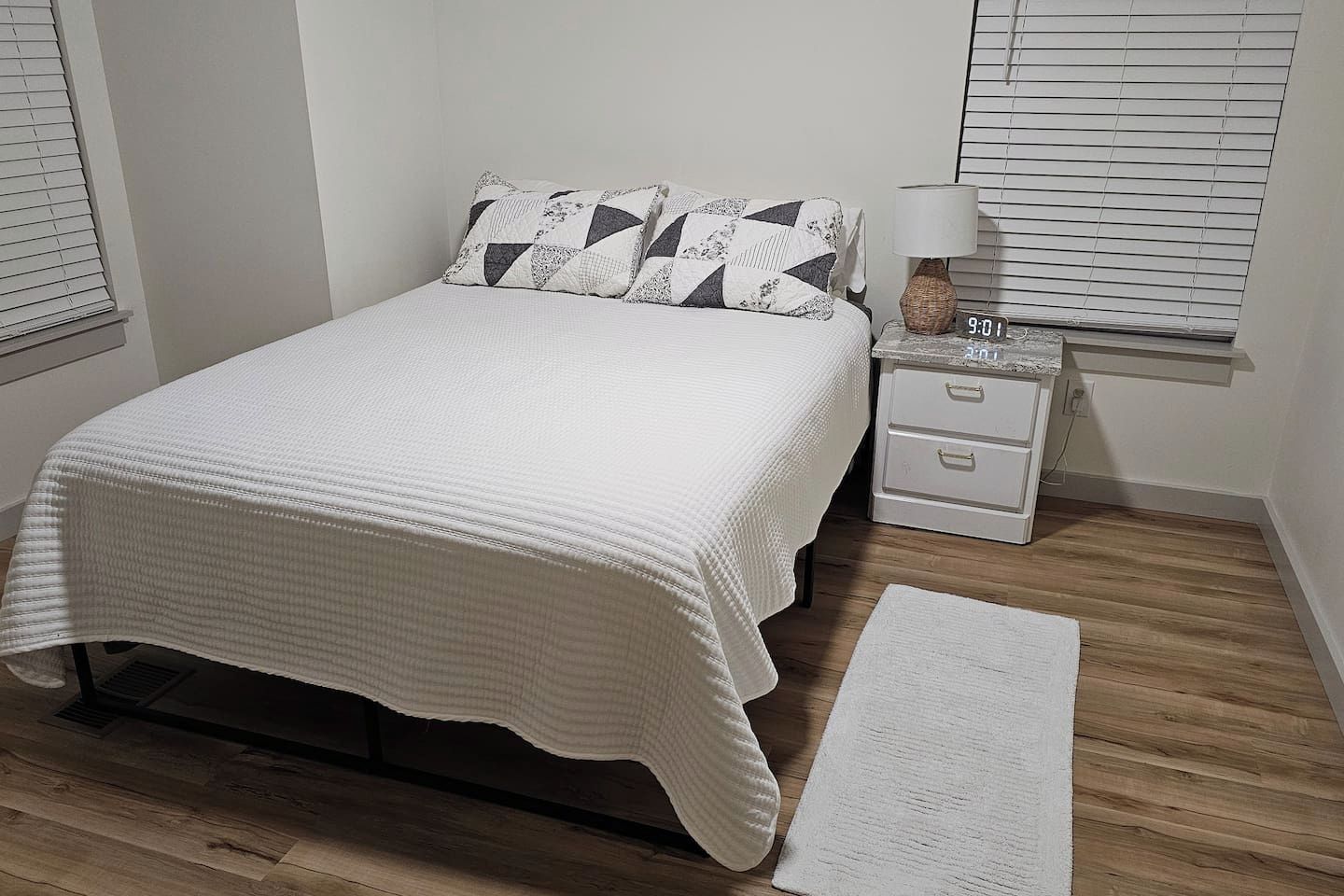 Un dormitorio con una cama, una mesita de noche con lámpara, una mesita auxiliar y un reloj digital.