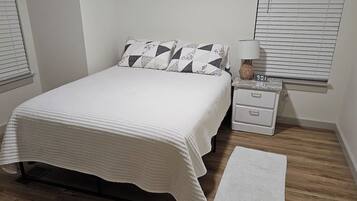 Un dormitorio con una cama, una mesita de noche con lámpara, una mesita auxiliar y un reloj digital.