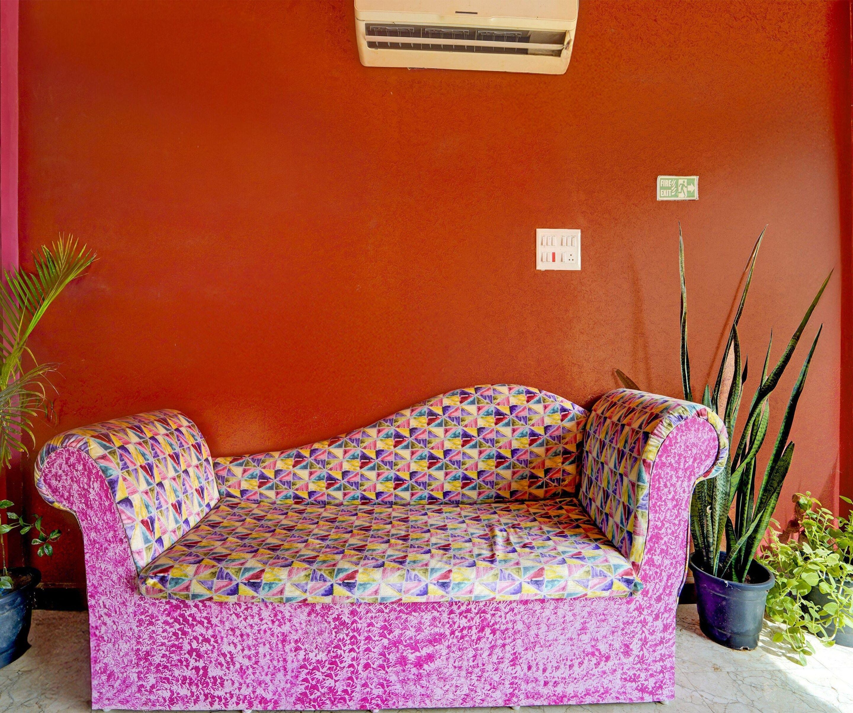 Un sofá colorido con un estampado geométrico, ubicado frente a una pared roja con un letrero de salida de incendios e interruptores eléctricos visibles.