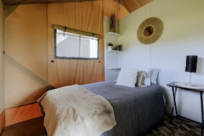 A bedroom with a bed, a bedside table, a lamp, a window, and a wall-mounted shelf.