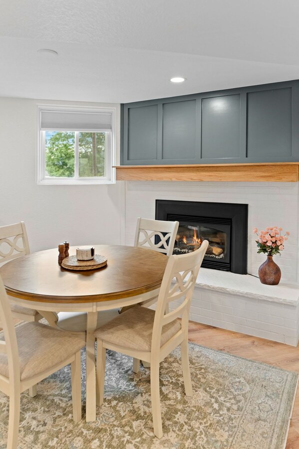 A dining area with a round wooden table, white chairs, a fireplace, and a window with a view of greenery.