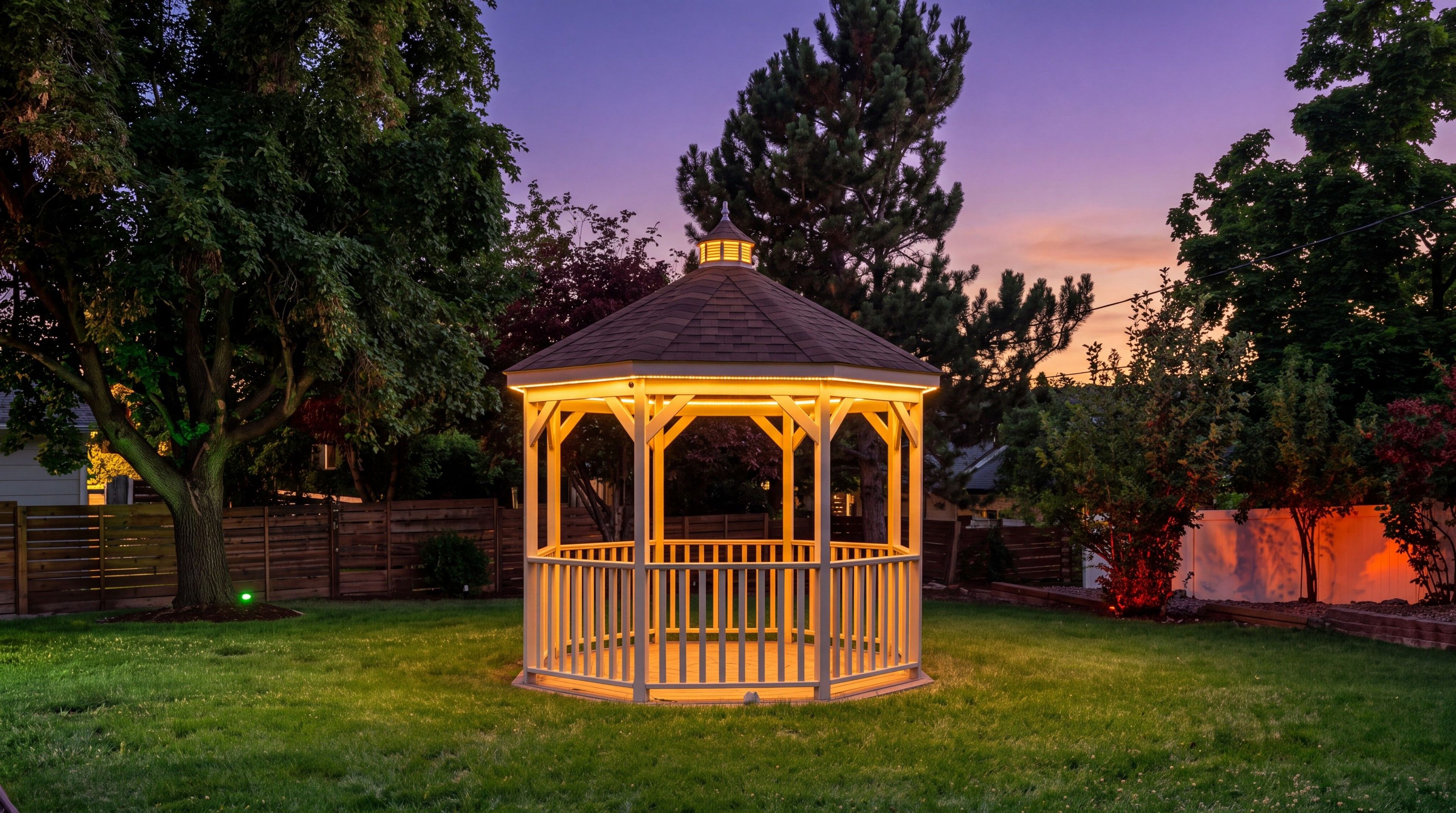 A gazebo with a hexagonal shape and a peaked roof, surrounded by a grassy yard and trees.