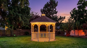 A gazebo with a hexagonal shape and a peaked roof, surrounded by a grassy yard and trees.
