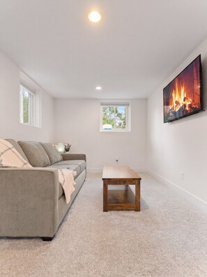 A room with a grey sofa, a wooden coffee table, a fireplace with a TV, and two windows.