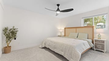 Un dormitorio con una cama, un ventilador de techo, una ventana y una mesita de noche.