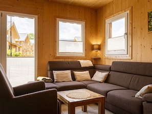 A cozy room with a brown sofa, a wooden table, and a lamp. There are large windows with blinds, and a view of a building outside. - One-bedroom Cabin (Sankt Georgen am Kreischberg)