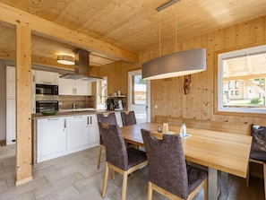 A wooden interior with a dining table and chairs, a kitchen with white cabinets, and a window with a view of a building outside. - One-bedroom Cabin (Sankt Georgen am Kreischberg)