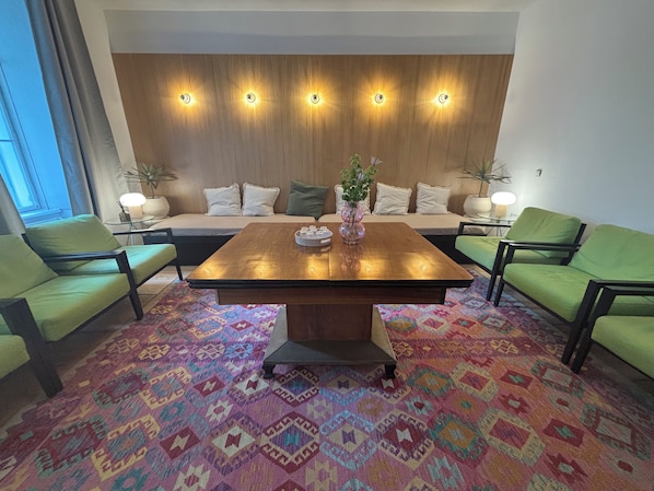Ein Zimmer mit einem Holztisch, grünen Sesseln, einem gemusterten Teppich und einer Wand mit Einbauleuchten.