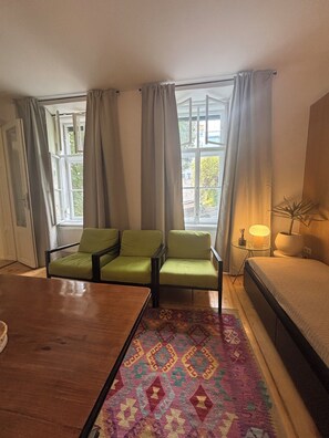 Ein Zimmer mit einem Holztisch, drei grünen Stühlen, einem gemusterten Teppich, einem Bett und einem Fenster mit Vorhängen.