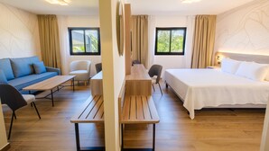 Quarto de hotel com cama, sofá, escrivaninha e cadeira. Janelas com cortinas, piso de madeira e parede com padrão de folhas.