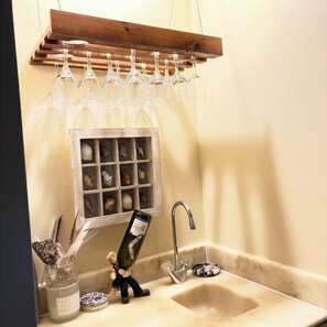 A bathroom with a wooden wine rack holding glasses, a decorative window with seashells, a sink with a faucet, and a figurine of a man with a bottle. - Marsh View Condo Steps to Beach Boardwalk - Pet Friendly (Saint Helena Island)