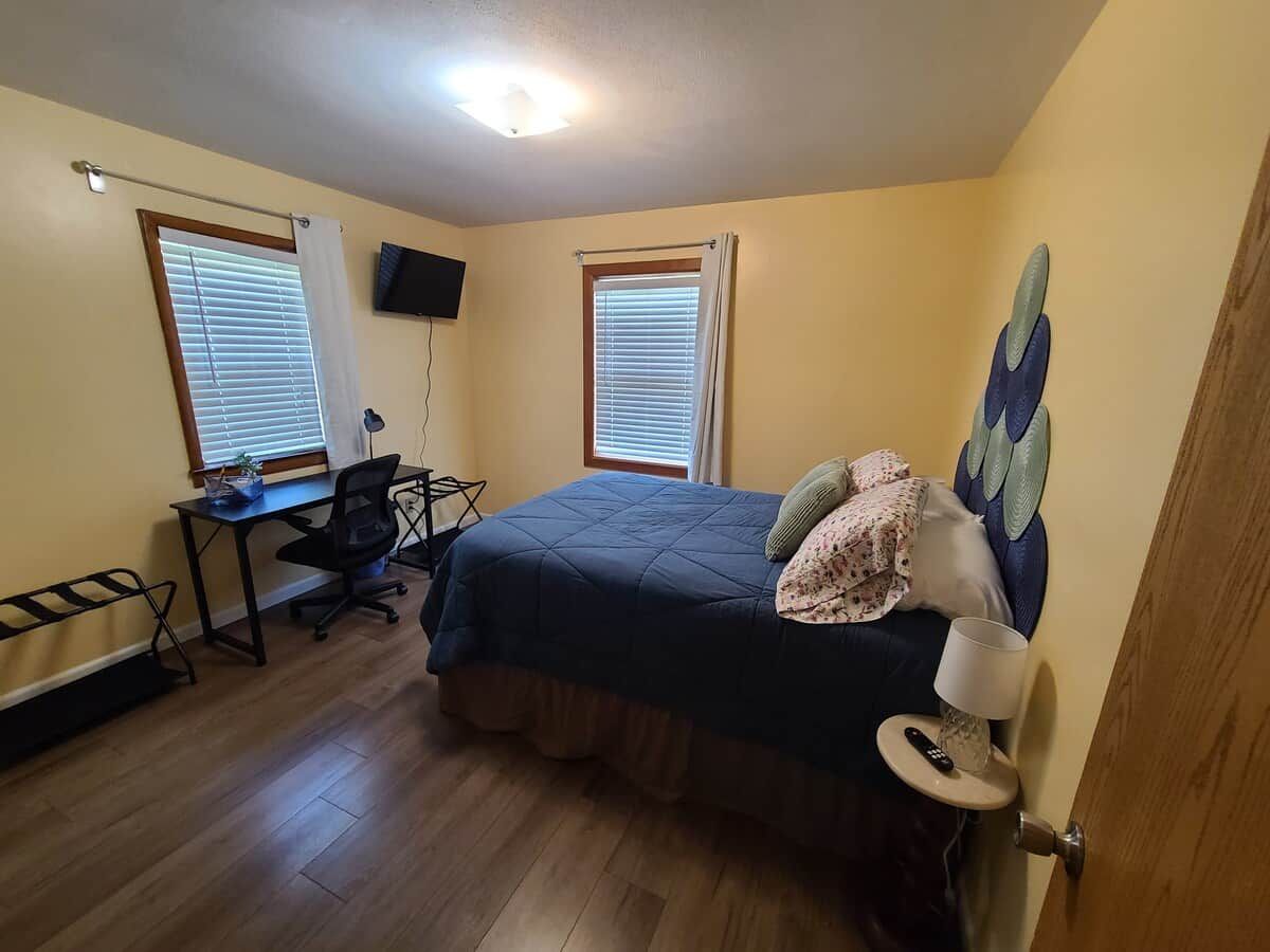 Quarto com cama, escrivaninha, cadeira e duas janelas com persianas.