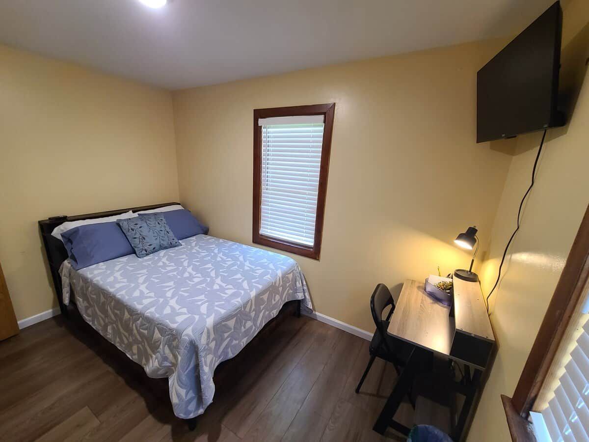 A bedroom with a bed, a desk, a chair, a window with blinds, and a wall-mounted TV.