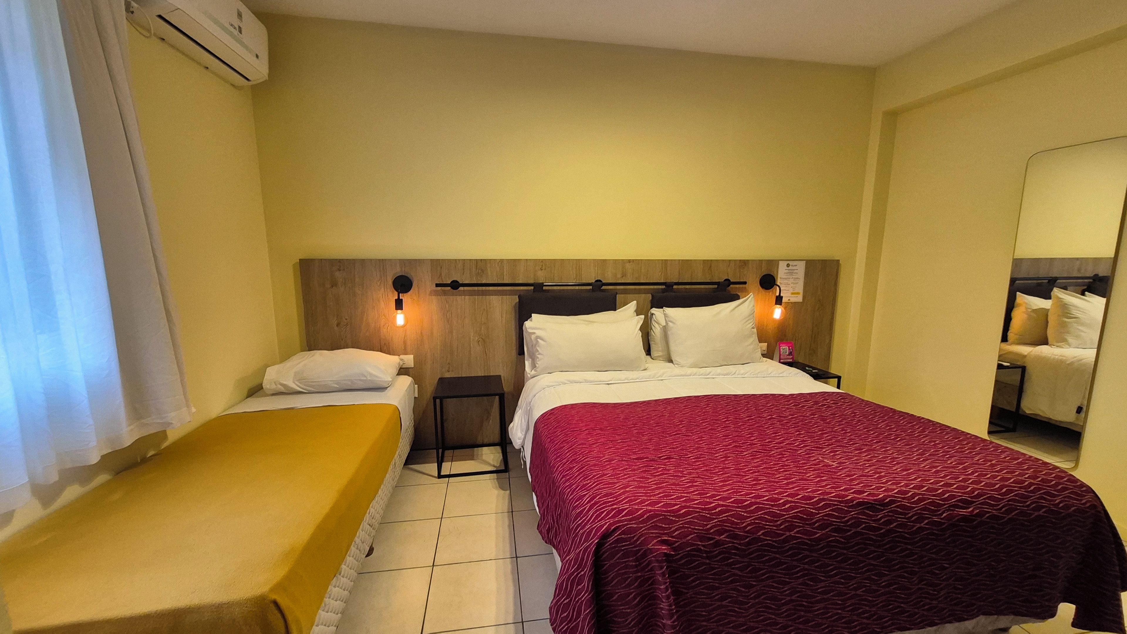 Hotellihuone, jossa on yksi sänky, pienempi sänky, yöpöytä, peili ja seinälle asennettu ilmastointilaite.