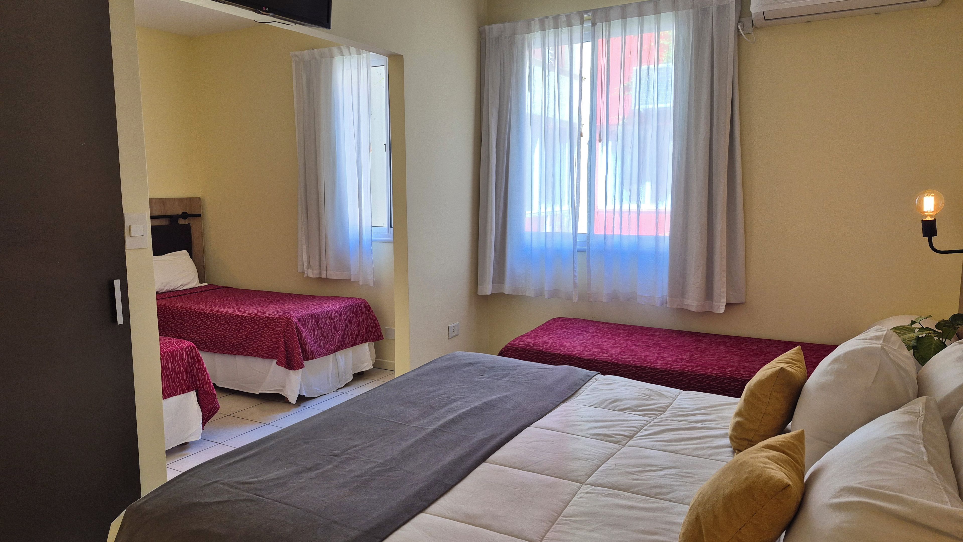 A room with two single beds, a larger bed, a bedside table, a lamp, and a window with curtains.