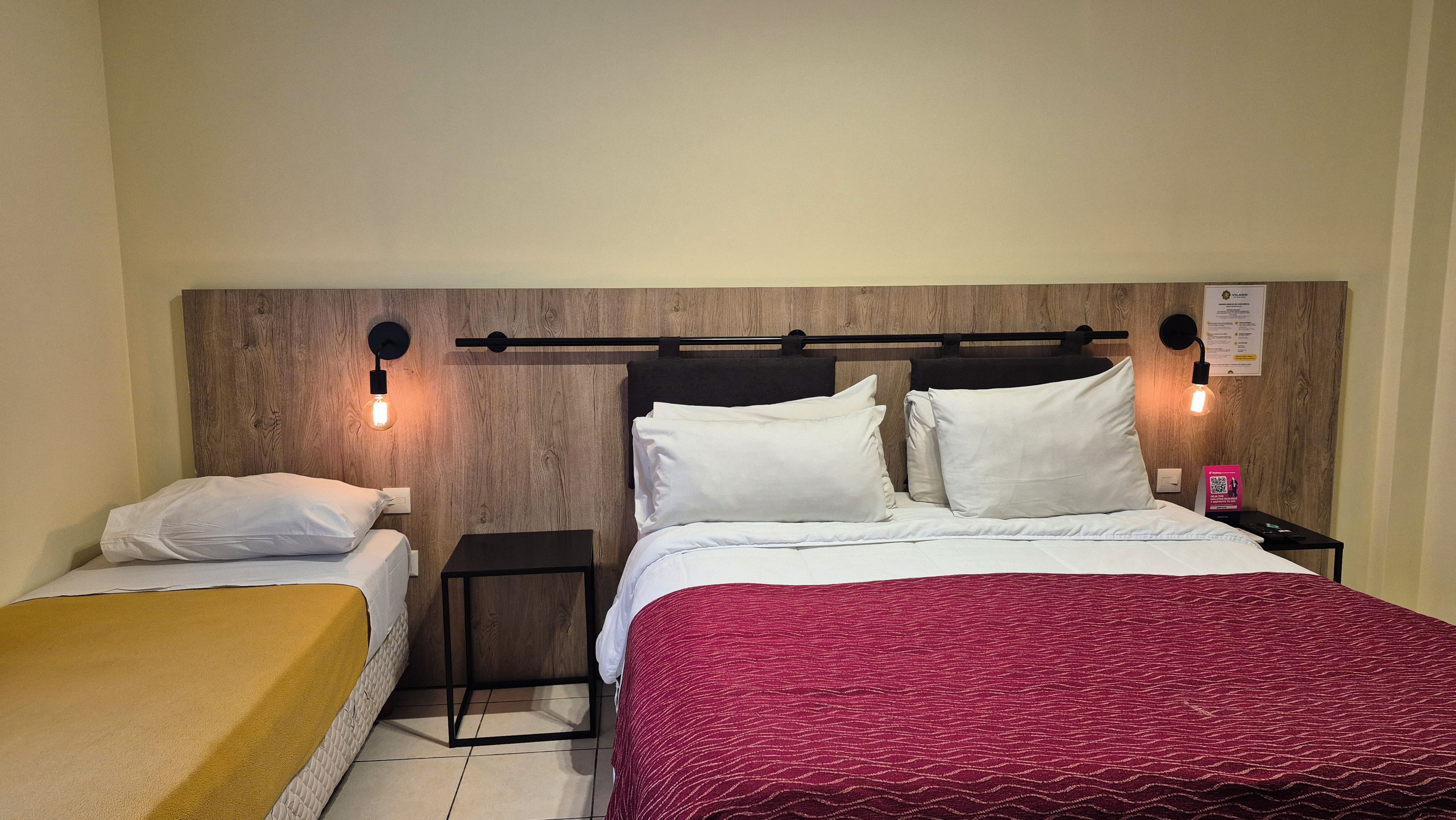 Habitación de hotel con cabecera de madera, dos mesitas de noche, cama con ropa blanca y una manta roja estampada, y lámparas de lectura fijadas a la pared.