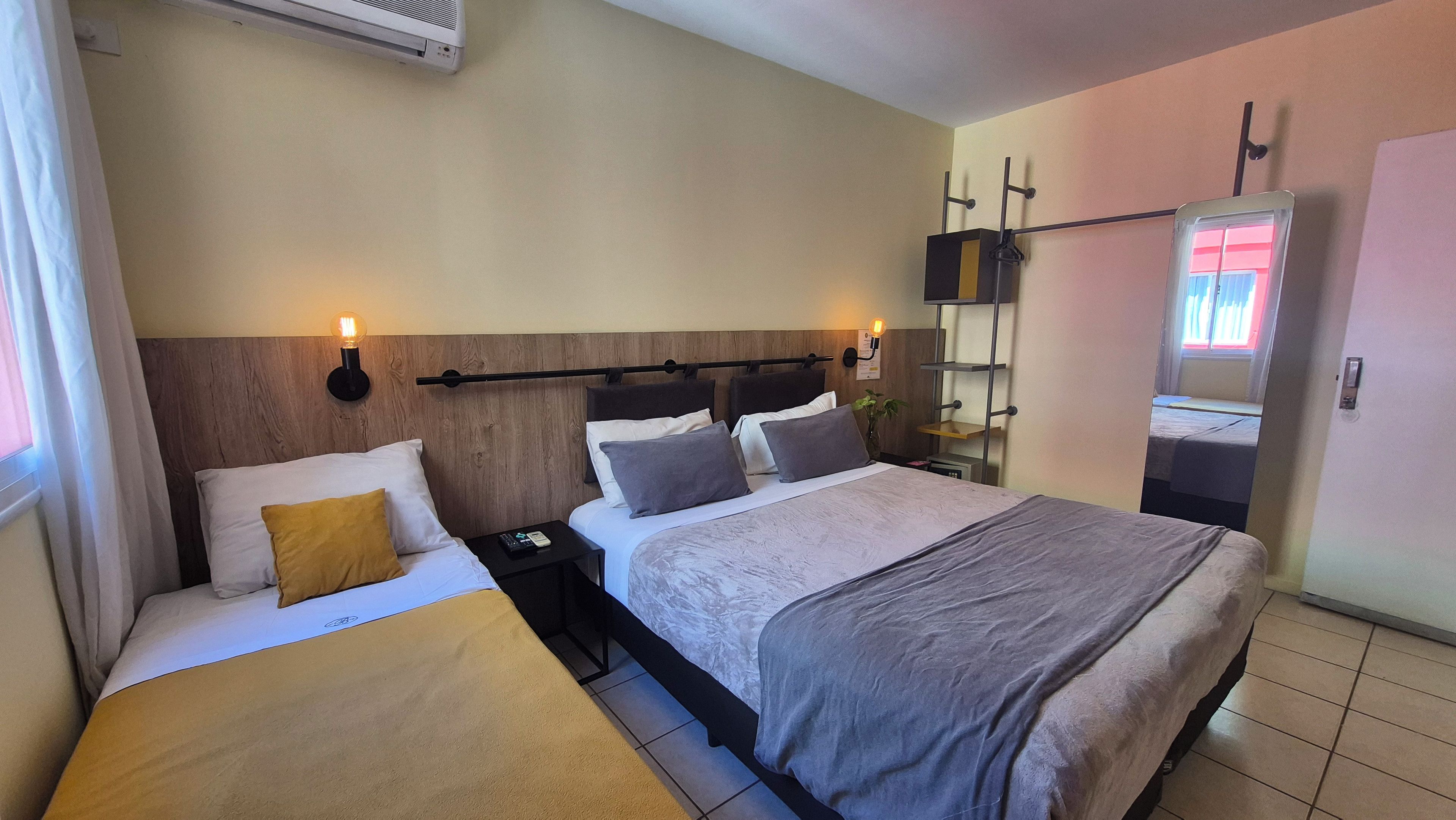 Habitación de hotel con cama, mesitas de noche, aire acondicionado de pared y un espejo sobre la puerta.