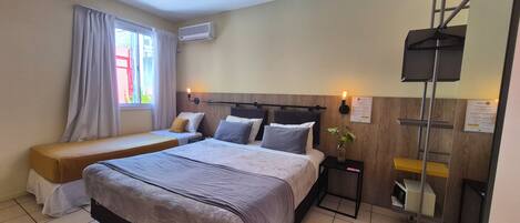 A hotel room with a double bed, a single bed, a bedside table with a lamp, an air conditioning unit, a window with curtains, and a wooden wall panel.