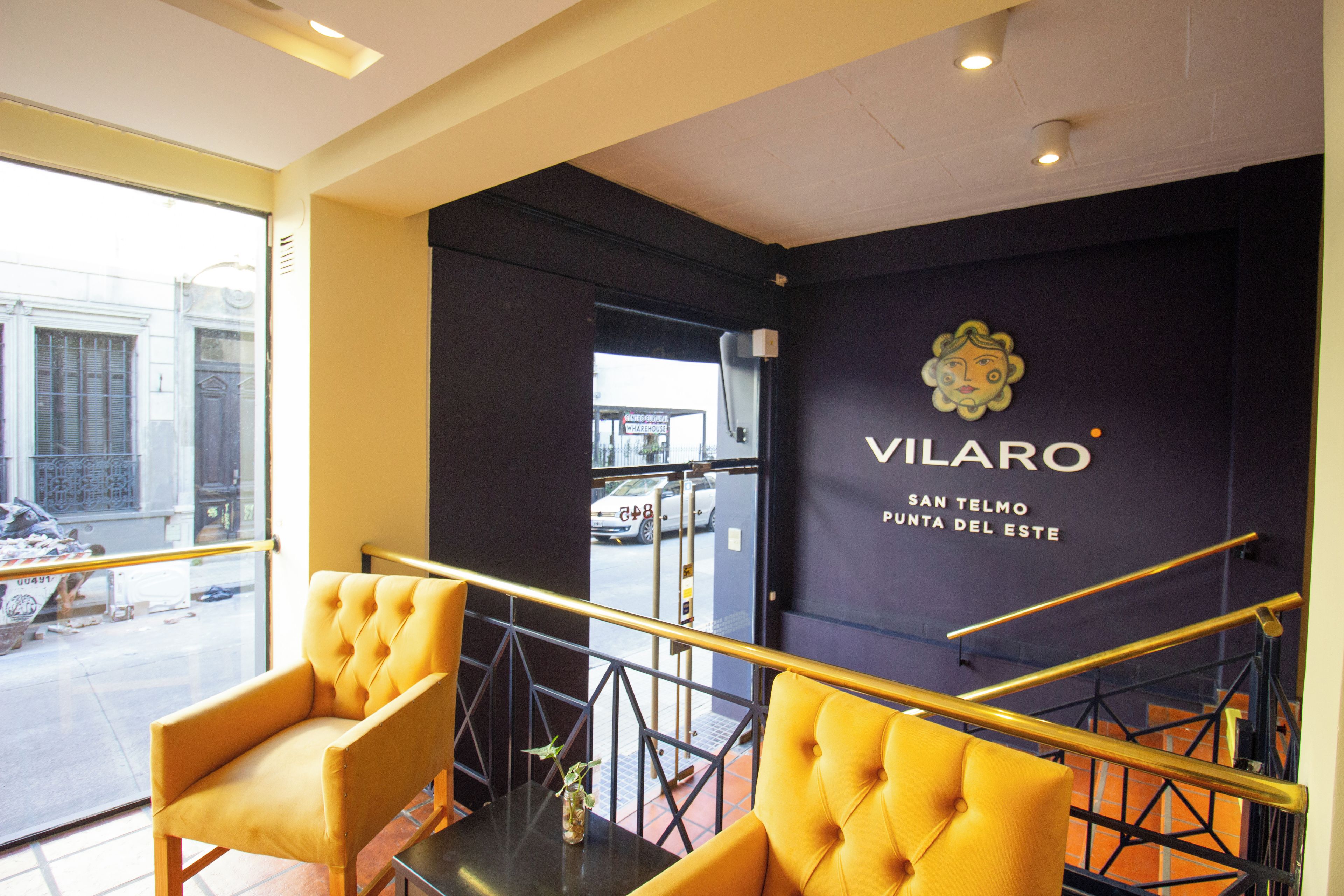 Interior de un hotel con una pared negra que muestra el nombre 'VILARO' y un cartel que dice 'SAN TELMO'. Se ven sillas amarillas y una mesita con un florero.