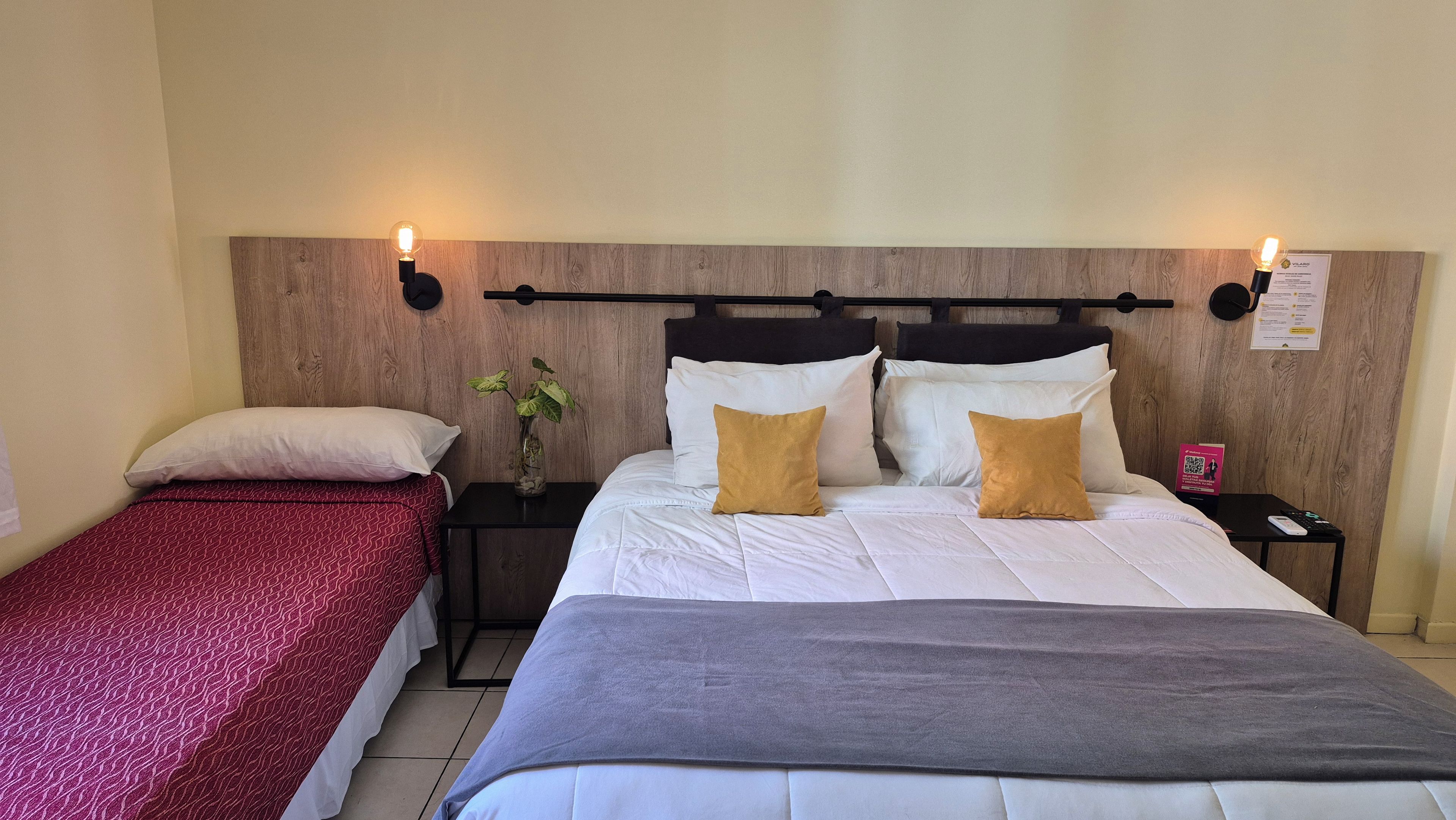 Habitación de hotel con cama doble, cama individual, mesitas de noche, un reloj, una planta y una lámpara fijada a la pared.