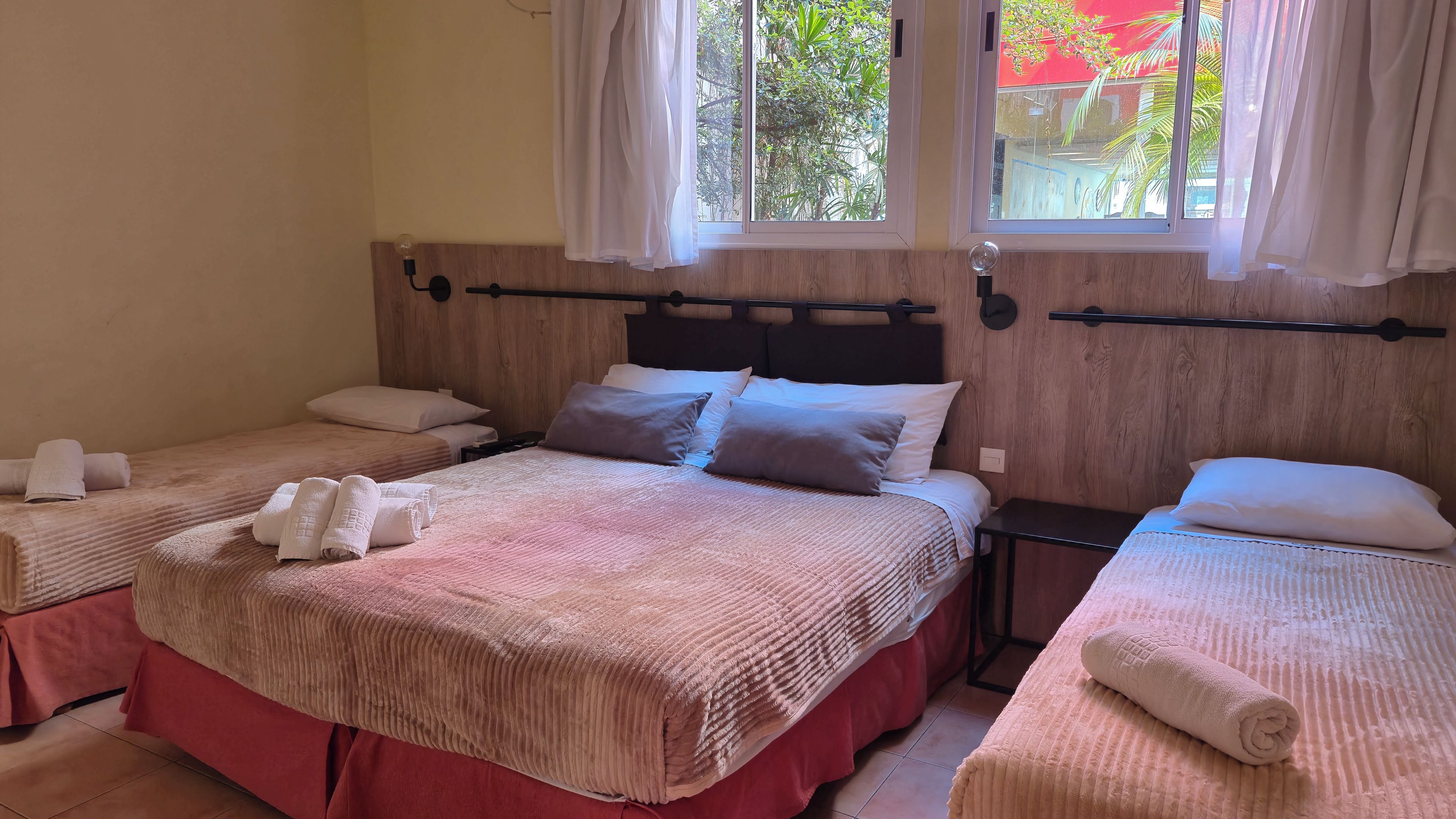 A hotel room with two beds, each with a headboard, pillows, and rolled towels. There are windows with curtains, and the room has a wooden headboard.