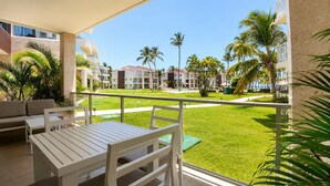 A balcony with a white table and chairs, overlooking a well-maintained lawn with palm trees and a building complex in the background. - Wake Up to the Ocean: 3-Bedroom Palmera · Beach & Pool (Punta Cana)
