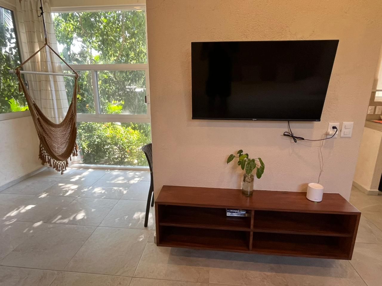 A living room with a flat-screen TV mounted on the wall, a wooden entertainment center, a hanging hammock, and a potted plant.