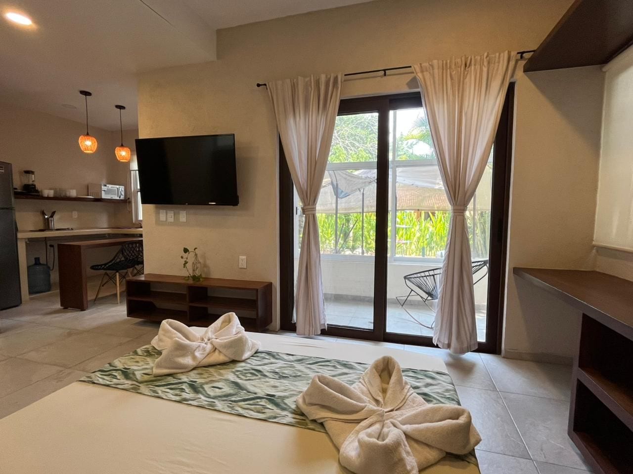 A hotel room with a bed, a TV, a kitchenette, and a sliding glass door to the outside.