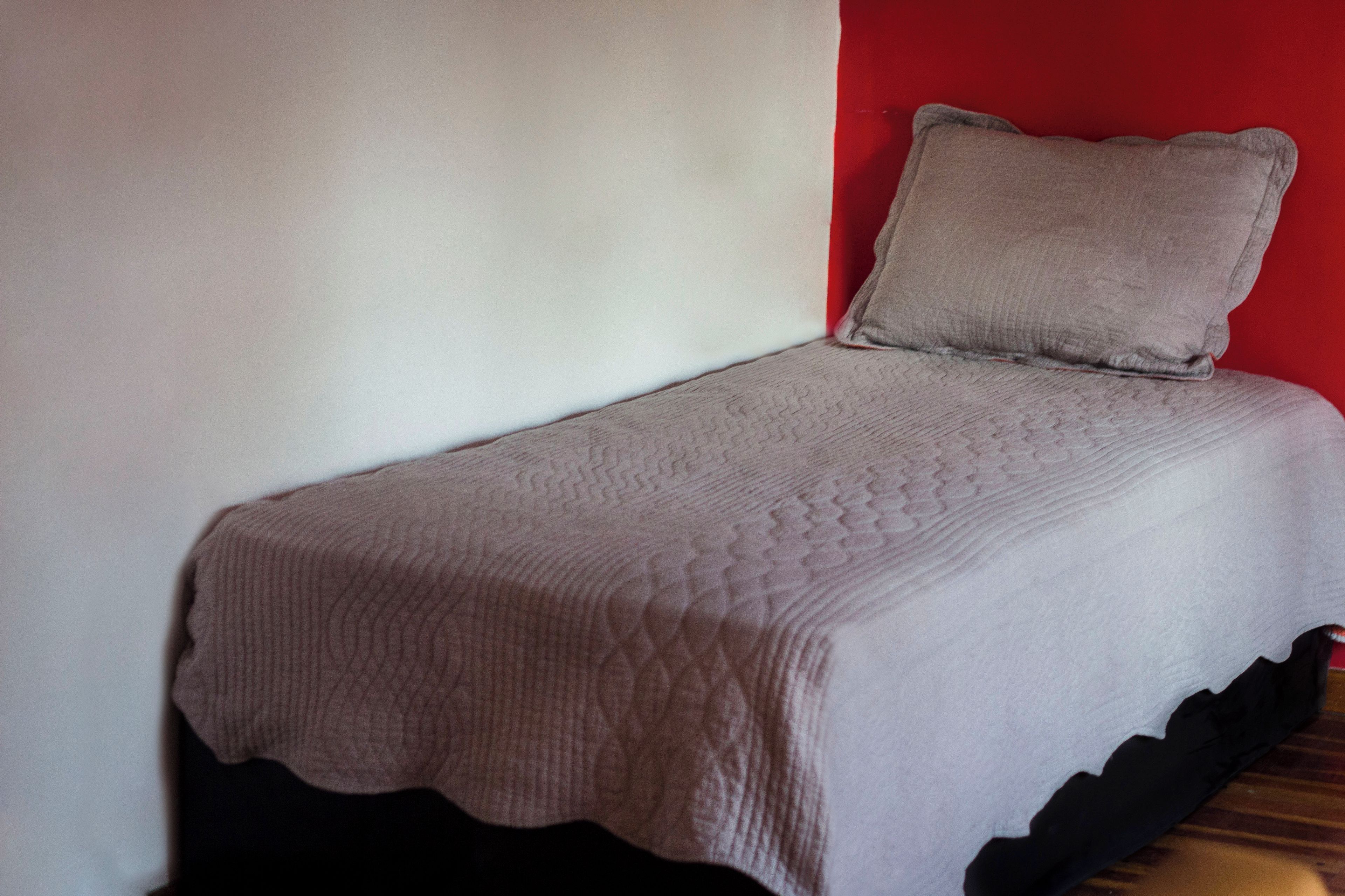 A single bed with a quilted bedspread and a pillow against a wall with a red accent.