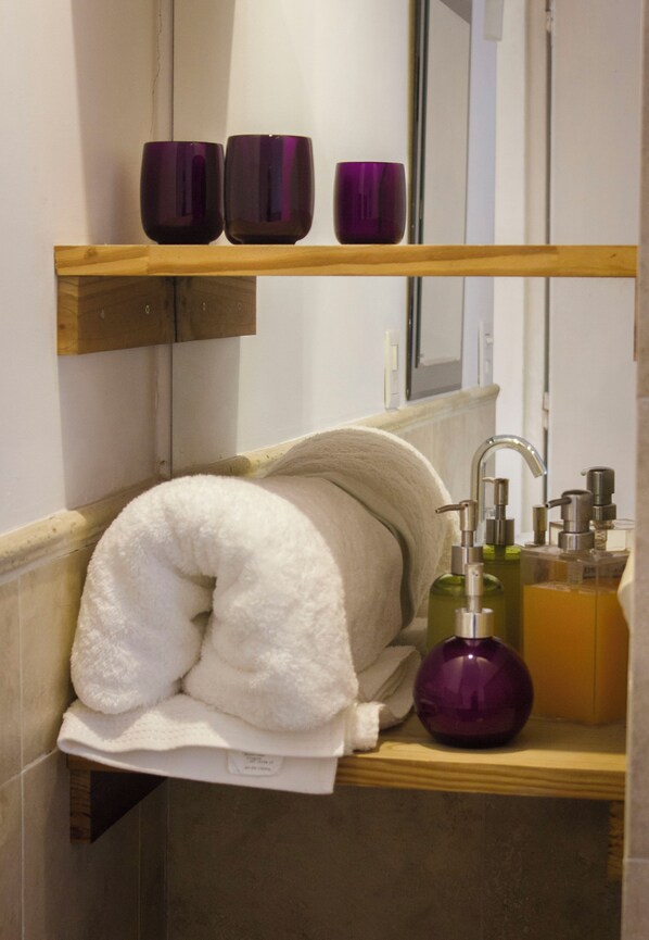 A bathroom with a towel rack holding a white towel, a wooden shelf with purple bottles, and a mirror above.