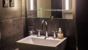 Un baño con un espejo rectangular, un lavamanos, un grifo, un dispensador de jabón y una copa de vidrio.