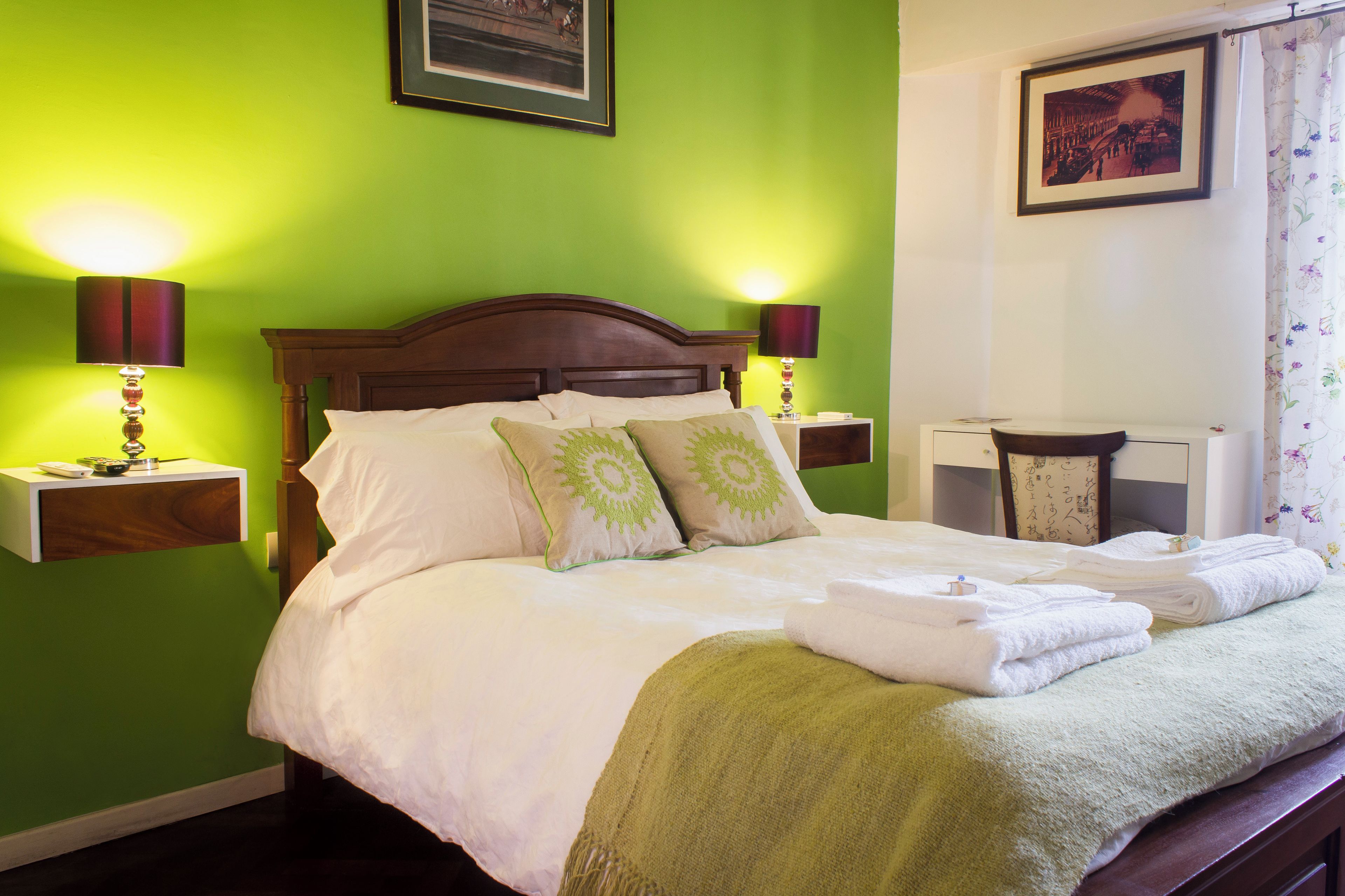 Una cama bien hecha con sábanas blancas y almohadas verdes, dos lámparas de noche y un cuadro colgado encima de la cama.
